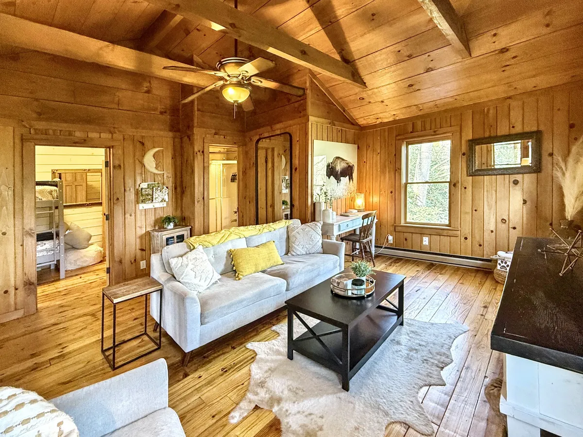 An inviting interior scene featuring a cozy sofa with yellow pillows, warm wooden walls and ceiling, a black coffee table, and a desk adorned with creative decorative items. The setup provides both comfort and a dedicated workspace, bathed in natural light.