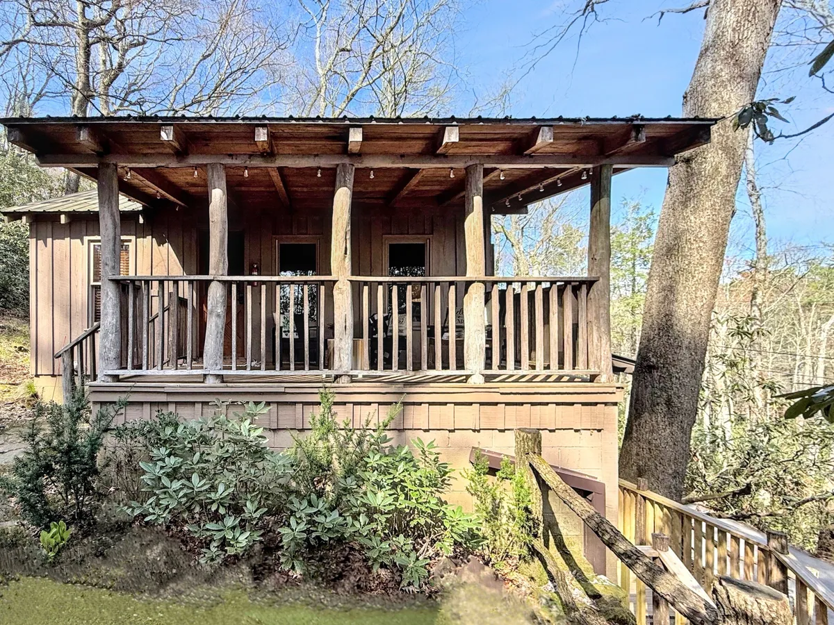 A wide wooden porch stretches across the home's rustic facade, facing lush greenery. The porch is shown as a versatile space for relaxing, entertaining guests, and soaking in views of sunrise or the fire pit nearby.