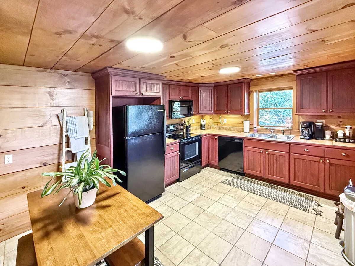 A charming and fully equipped kitchen with warm-toned cabinets, black appliances, a wooden table, and a window offering a pleasant view. The space feels both practical and inviting, ideal for preparing meals or gathering for cozy indoor dining.