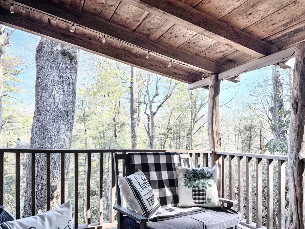 The hero image showcases the inviting exterior porch of a restored 1930's cabin, complete with a classic wooden railing, plush plaid cushions, and decorative pillows, all set against a backdrop of lush Highlands greenery. The setting is ideal for relaxing with a book, sipping coffee at sunrise, or enjoying the tranquil mountain air.
