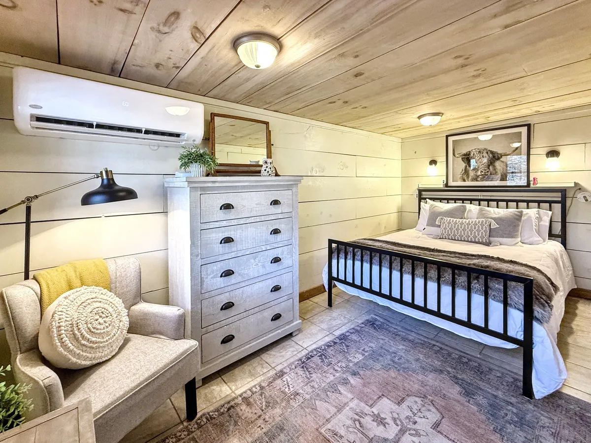 A tranquil bedroom scene with a bed dressed in soft linens, a cozy reading chair next to it, a dresser with decorative accents, and a framed picture on the wall. Gentle daylight filters through the window, enhancing the room’s peaceful, restorative quality ideal for restful nights or quiet moments spent indoors.