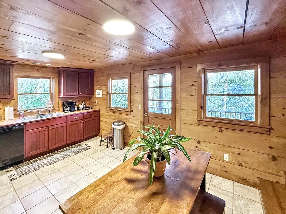 A homey kitchen and dining area with a central table, kitchen cabinets, a sink, large windows, and a potted plant. The space is filled with natural light and offers views of the surrounding nature, creating an inviting environment for meals, coffee breaks, or casual laptop sessions.