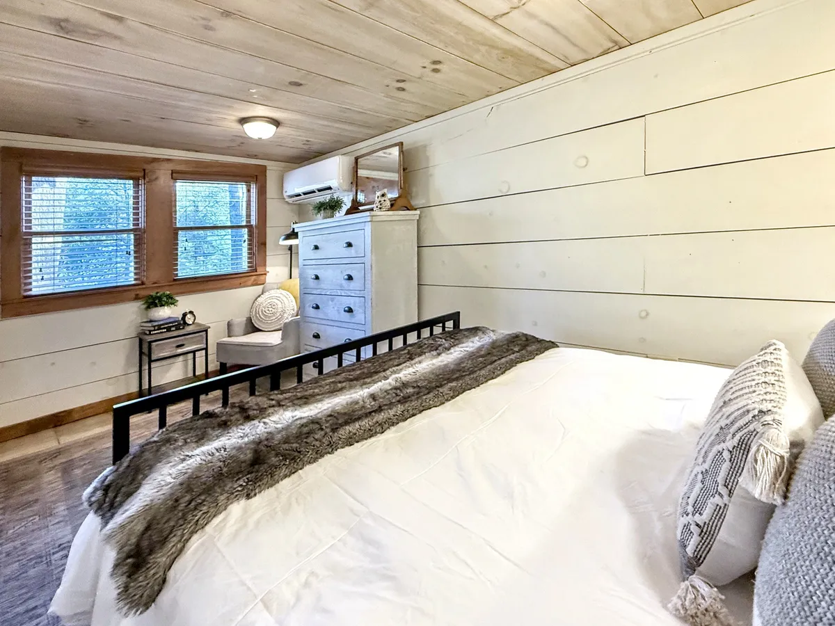 A serene bedroom scene featuring a bed with a fur throw, an armchair, dresser with mirror, and multiple windows letting in natural light. The coziness of the bedding hints at winter warmth, while the abundant daylight and nature views evoke the lush, vibrant seasons.