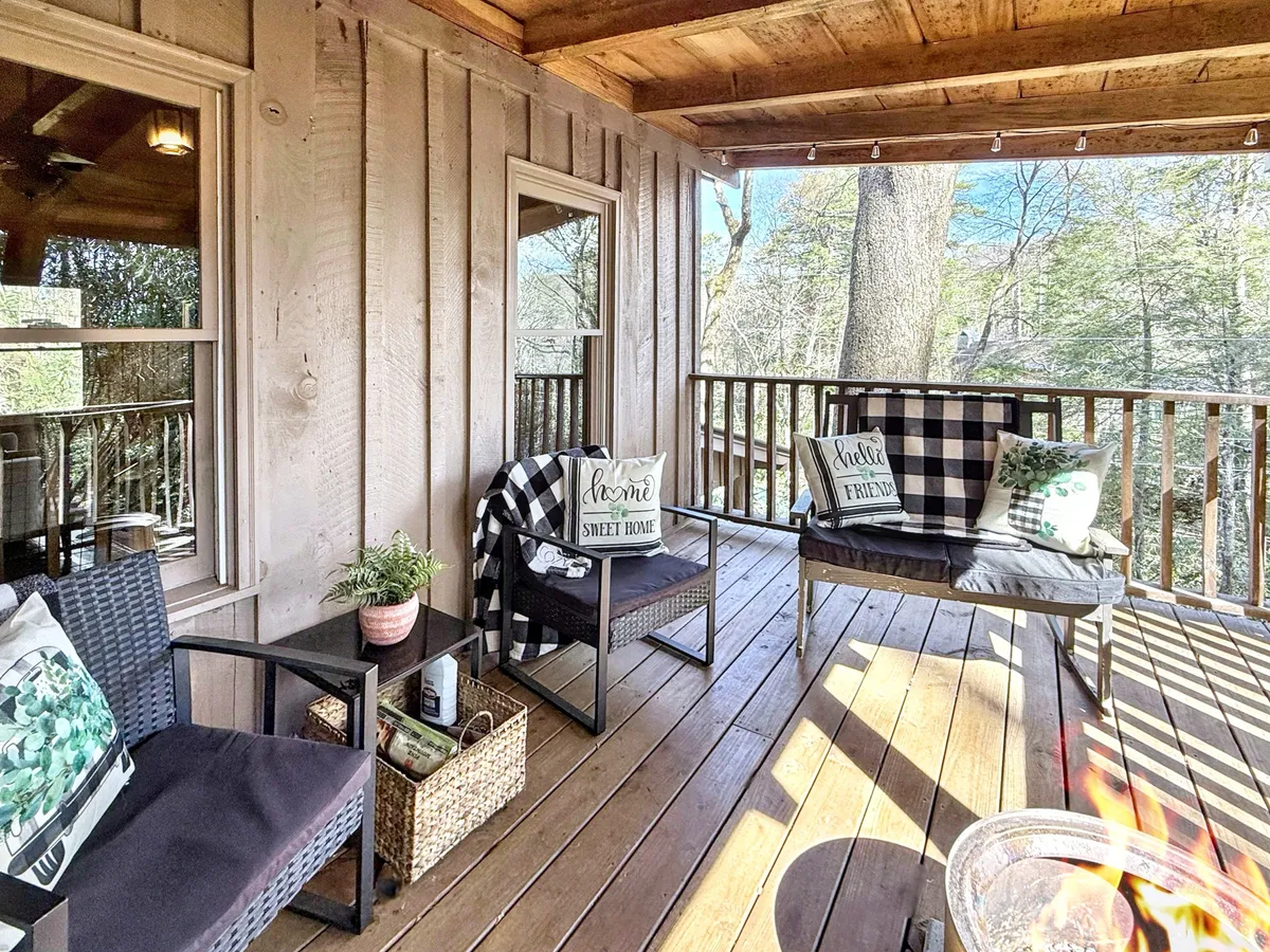 A bright, cozy living area featuring comfortable seating with decorative pillows arranged around a wooden railing. Large windows frame lush, natural surroundings, allowing woodland views to filter in and filling the space with a serene, adaptable atmosphere. Subtle touches like blankets and textured throws hint at comfort for every season.