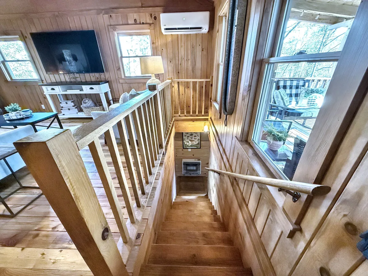 A bright, welcoming interior view showcasing a wooden staircase, light fixture, wall-mounted television, lamp, and a comfortable seating area. The cozy arrangement invites guests to relax, entertain, or enjoy a cup of coffee, all set against a backdrop of rich woodwork.