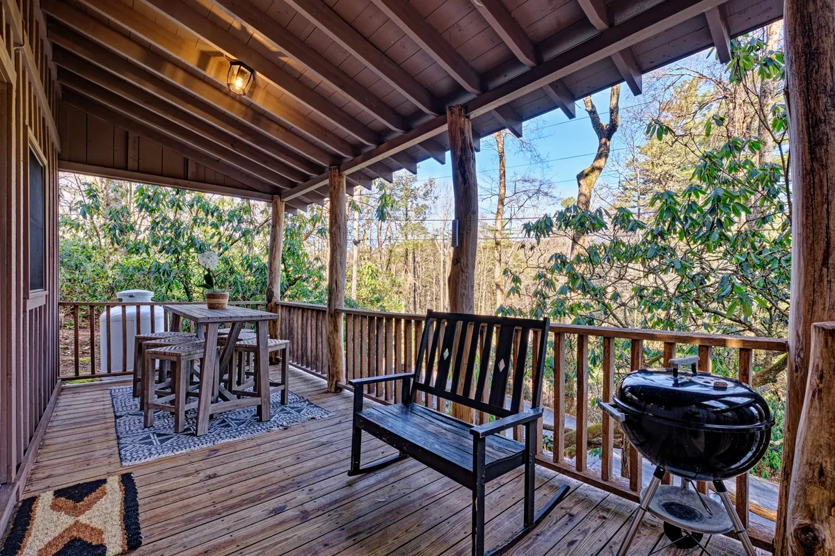 A charming porch scene featuring a wooden table set for dining, a classic black grill ready for barbecuing, and a rocking chair. The porch is surrounded by lush greenery, providing a peaceful and inviting atmosphere perfect for alfresco meals and relaxed conversations.