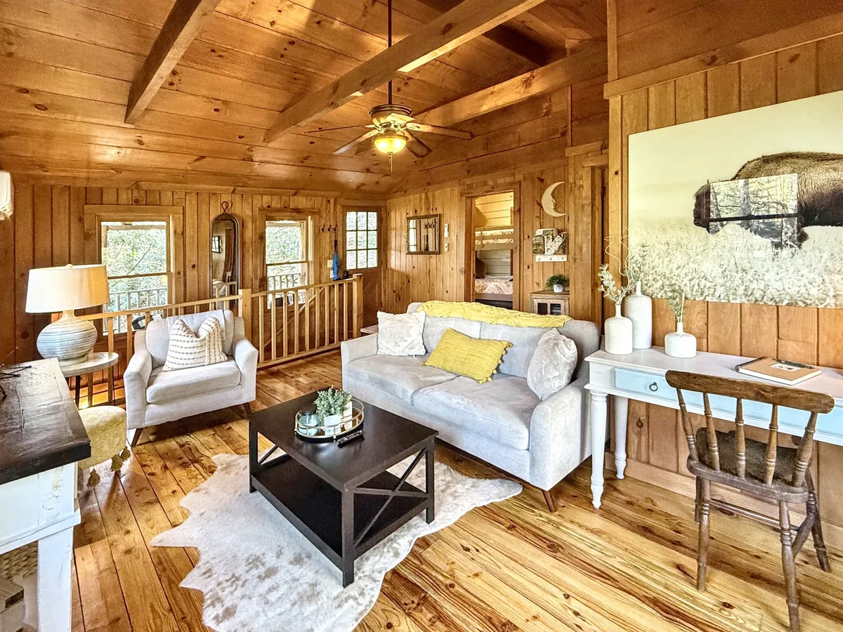 A warm and welcoming living room setting with a sofa, coffee table, and a lamp casting mood lighting. The room invites relaxation, reading, and socializing, with artwork on the wall and a window offering a glimpse of the outdoors. The ambiance is perfect for quiet mornings or gathering with friends regardless of the season.