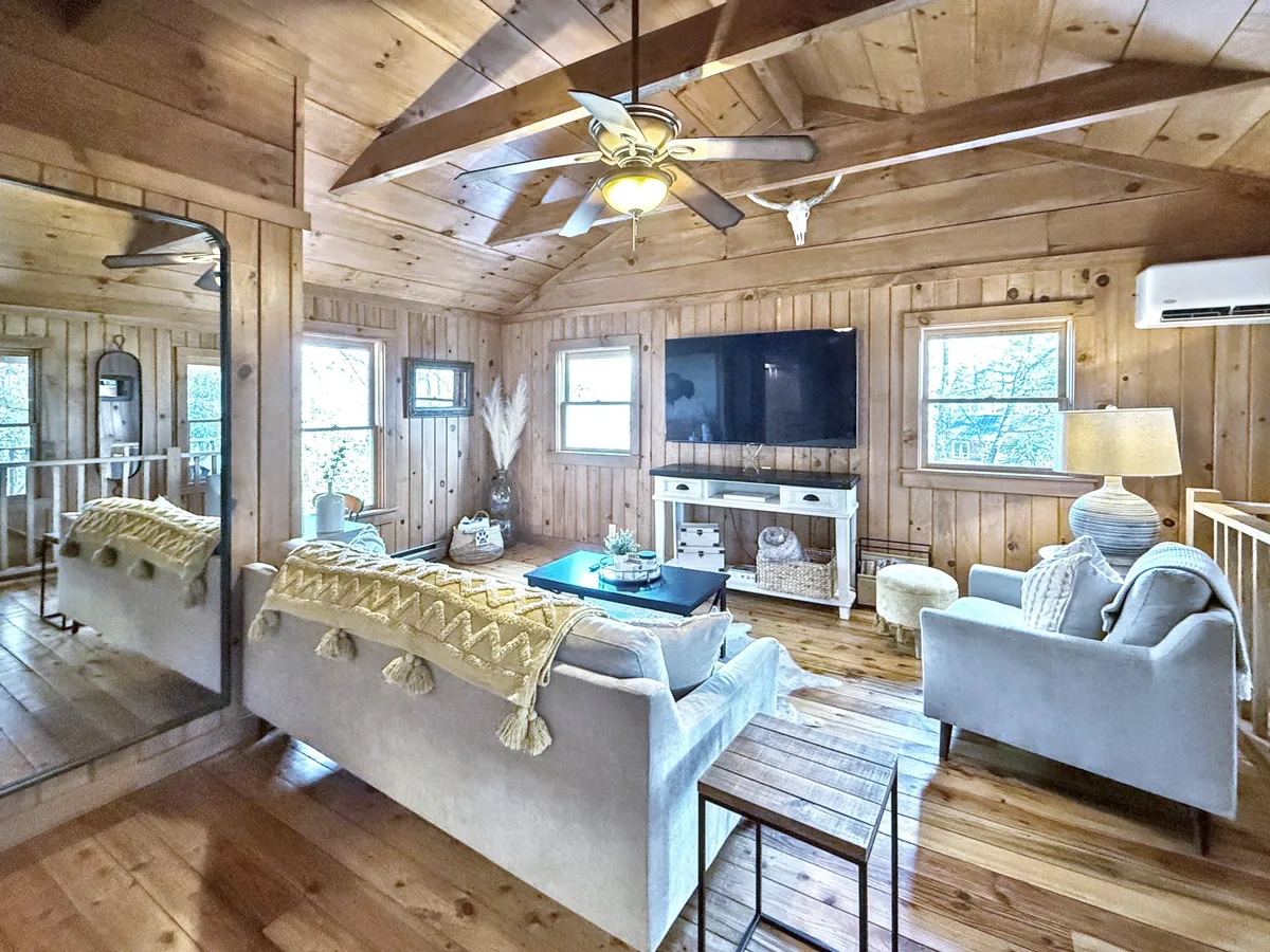 A warm and inviting living room featuring a comfortable sofa with a decorative throw, a central coffee table, TV, and beautiful wood-paneled ceiling and walls. A softly glowing lamp adds a cozy ambiance, highlighting the hardwood floors. The setting blends historic charm with contemporary comfort, echoing the spirit of the lovingly restored cabin.
